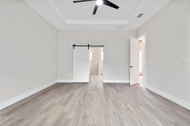 empty room featuring ceiling fan, a barn door, a raised ceiling, and light hardwood / wood-style flooring