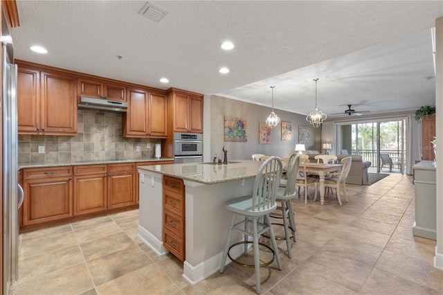 kitchen with a kitchen bar, hanging light fixtures, light stone countertops, a kitchen island with sink, and backsplash