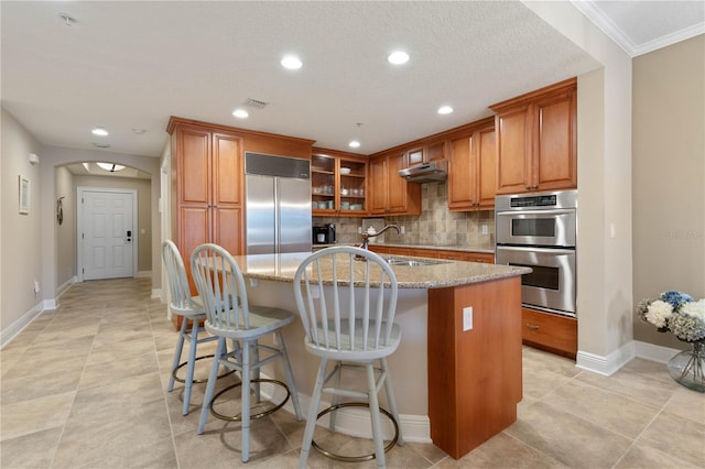 kitchen with appliances with stainless steel finishes, a breakfast bar, backsplash, light stone countertops, and a center island with sink