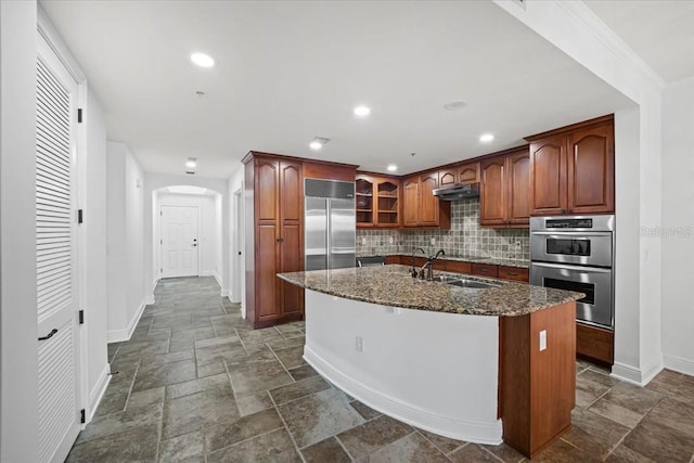 kitchen with sink, appliances with stainless steel finishes, an island with sink, dark stone counters, and decorative backsplash