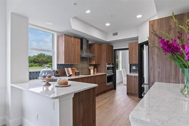 kitchen featuring wall chimney range hood, light hardwood / wood-style flooring, backsplash, stainless steel appliances, and kitchen peninsula