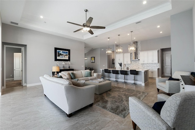 living room with sink, ceiling fan with notable chandelier, ornamental molding, and a raised ceiling