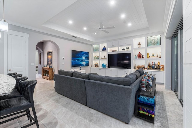 living room featuring arched walkways, built in features, a tray ceiling, and ornamental molding