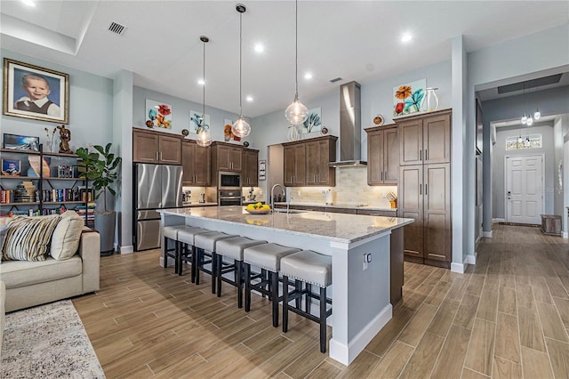 kitchen with pendant lighting, a breakfast bar, light stone countertops, a center island with sink, and wall chimney exhaust hood