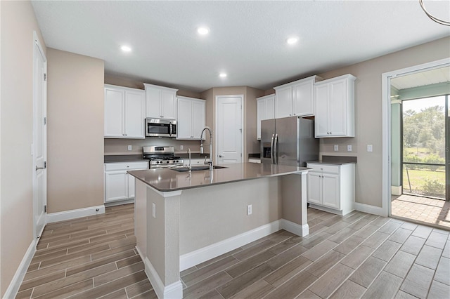 kitchen featuring dark countertops, an island with sink, stainless steel appliances, white cabinetry, and a sink