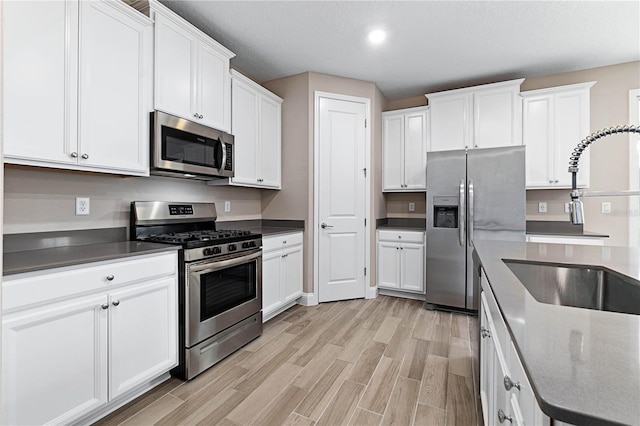 kitchen with stainless steel appliances, a sink, white cabinetry, light wood finished floors, and dark countertops