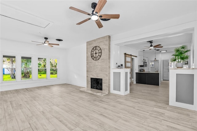unfurnished living room featuring a tile fireplace, a barn door, ceiling fan, and light wood-type flooring