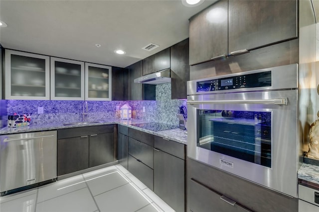 kitchen with visible vents, appliances with stainless steel finishes, glass insert cabinets, light stone countertops, and under cabinet range hood