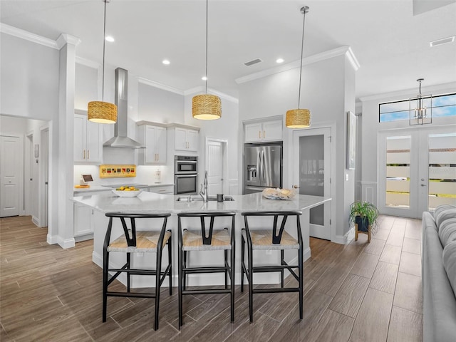 kitchen with white cabinets, appliances with stainless steel finishes, wall chimney range hood, a large island with sink, and pendant lighting