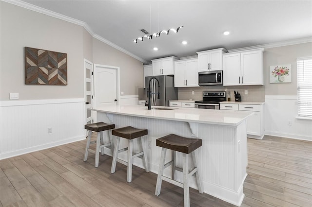 kitchen featuring light wood finished floors, an island with sink, appliances with stainless steel finishes, crown molding, and white cabinetry