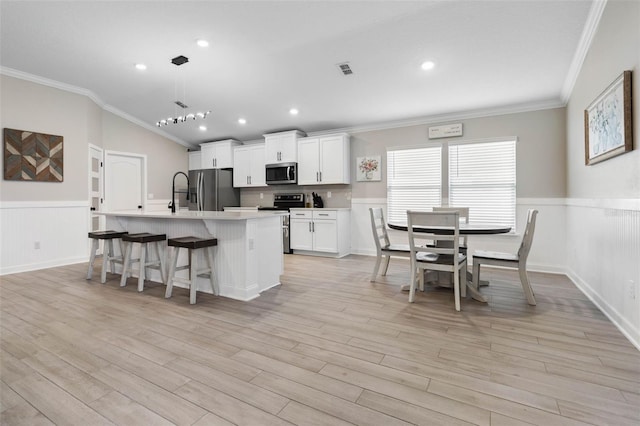 kitchen with light wood finished floors, wainscoting, a kitchen island with sink, stainless steel appliances, and light countertops