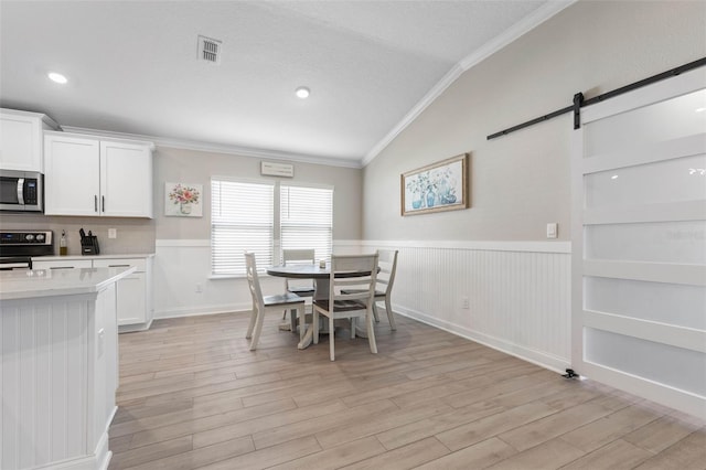 dining room with a barn door, wainscoting, lofted ceiling, light wood-style flooring, and ornamental molding