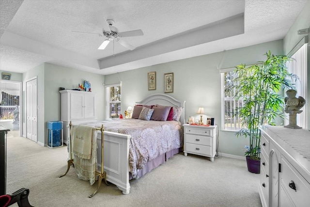 bedroom featuring light carpet, baseboards, ceiling fan, a tray ceiling, and a textured ceiling