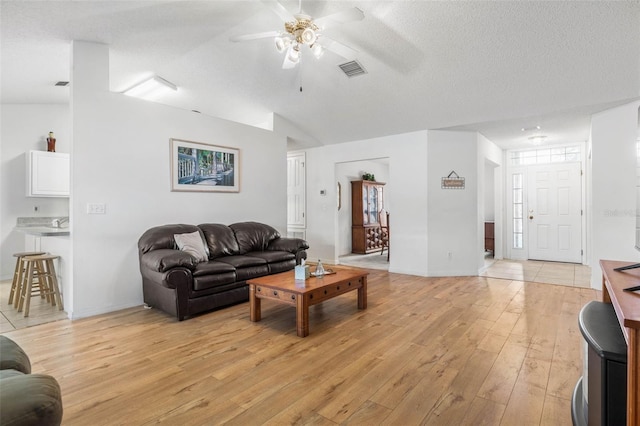 living room featuring ceiling fan, a textured ceiling, visible vents, vaulted ceiling, and light wood finished floors