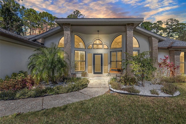 exterior entry at dusk with brick siding, a lawn, and stucco siding