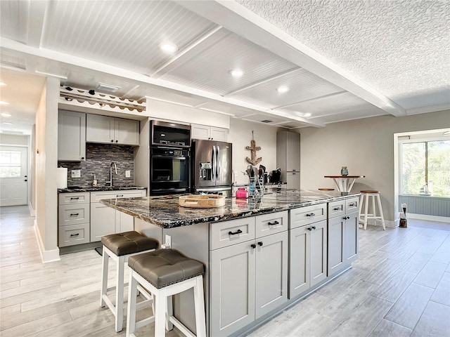 kitchen with stainless steel refrigerator with ice dispenser, light wood finished floors, gray cabinetry, an island with sink, and dark stone counters
