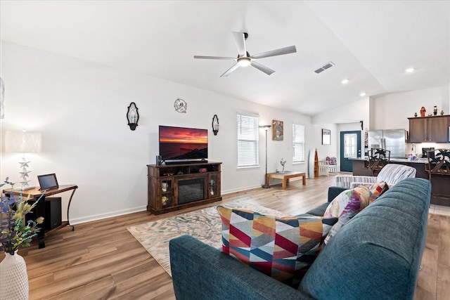 living room featuring visible vents, a glass covered fireplace, ceiling fan, light wood-style flooring, and vaulted ceiling