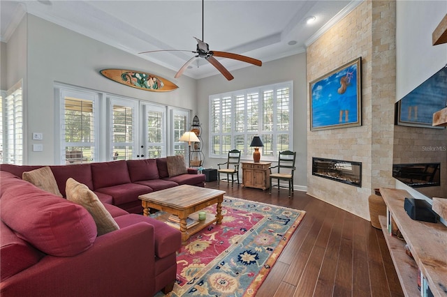 living room featuring dark wood-style floors, a raised ceiling, ornamental molding, a large fireplace, and ceiling fan