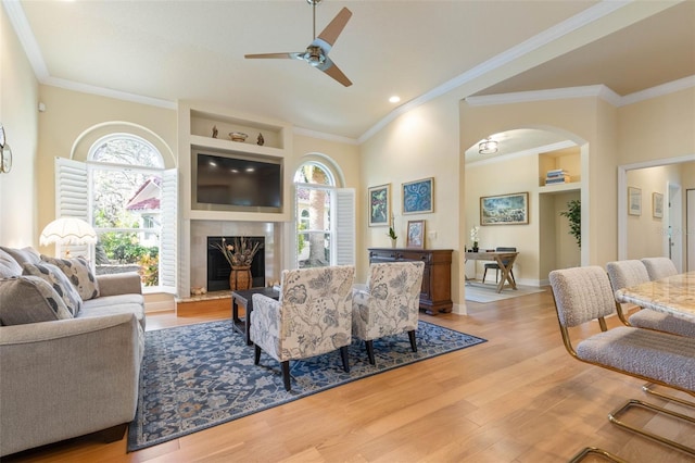 living room featuring a fireplace with raised hearth, arched walkways, wood finished floors, a ceiling fan, and ornamental molding