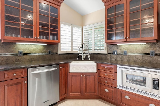 kitchen with stainless steel appliances, backsplash, glass insert cabinets, a sink, and dark stone countertops