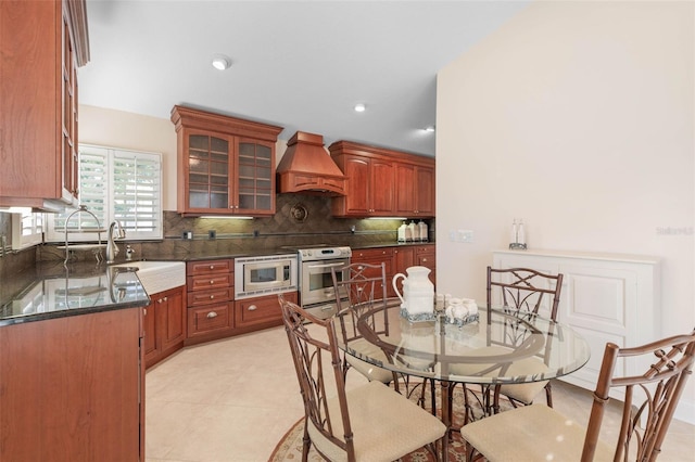 kitchen featuring tasteful backsplash, glass insert cabinets, appliances with stainless steel finishes, premium range hood, and a sink