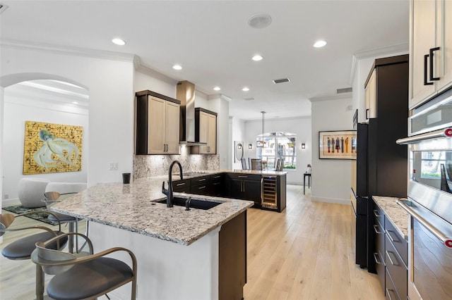 kitchen featuring backsplash, wall chimney range hood, wine cooler, a peninsula, and a sink