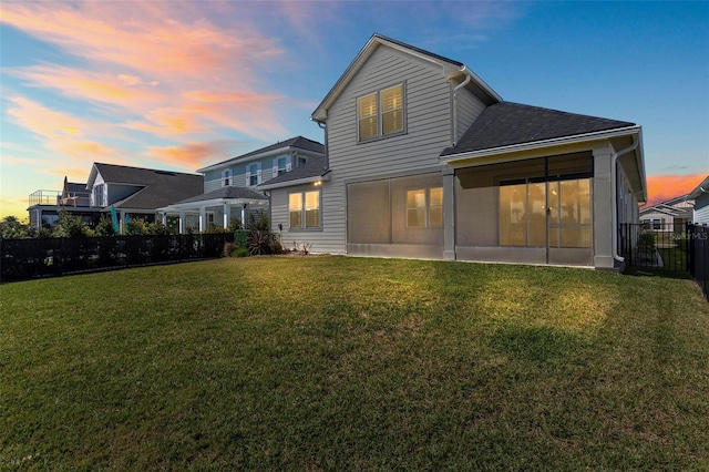 back of property at dusk featuring a lawn, a sunroom, roof with shingles, and fence