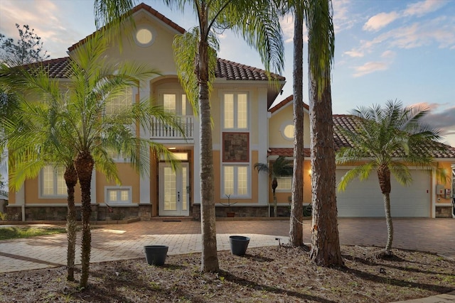 mediterranean / spanish home featuring decorative driveway, a tile roof, stucco siding, an attached garage, and a balcony