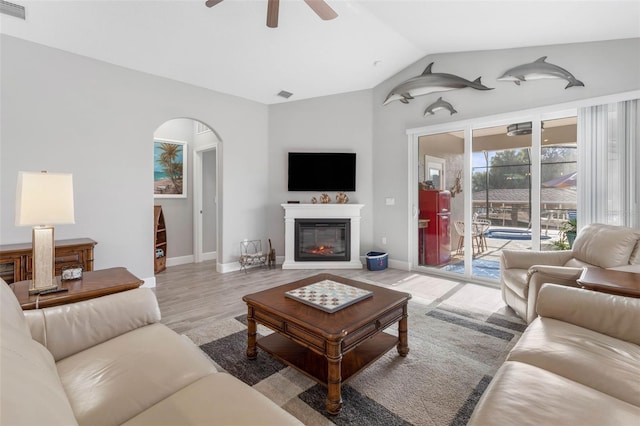 living room featuring arched walkways, visible vents, a ceiling fan, vaulted ceiling, and a glass covered fireplace