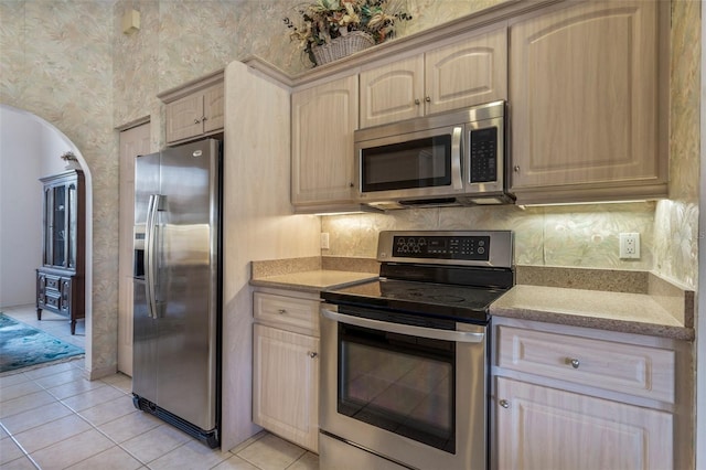 kitchen featuring stainless steel appliances, tasteful backsplash, light tile patterned flooring, light brown cabinets, and wallpapered walls