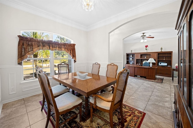 dining space with arched walkways, a decorative wall, a wainscoted wall, ceiling fan with notable chandelier, and crown molding