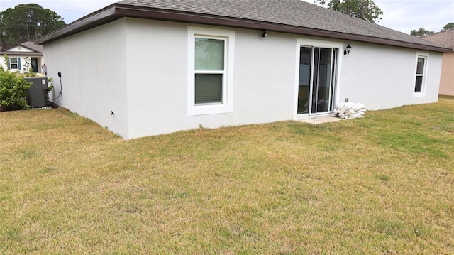 rear view of house with roof with shingles, central AC unit, a lawn, and stucco siding