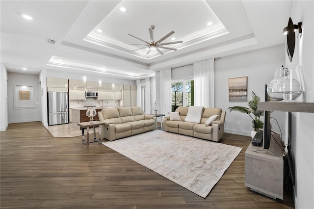 living area with a ceiling fan, visible vents, baseboards, dark wood-style floors, and a tray ceiling