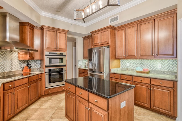 kitchen featuring brown cabinetry, wall chimney exhaust hood, a kitchen island, appliances with stainless steel finishes, and dark stone countertops