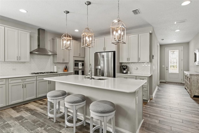 kitchen with stainless steel appliances, a sink, wood finished floors, wall chimney range hood, and a center island with sink