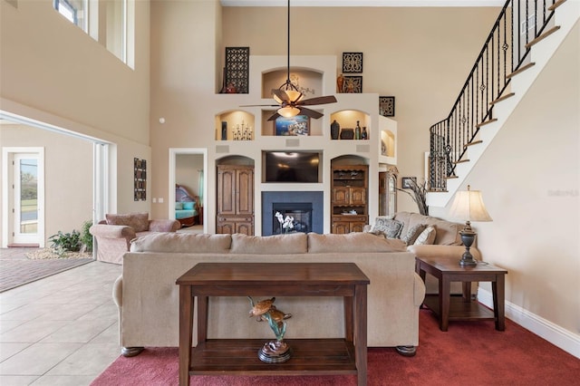 tiled living room featuring baseboards, stairs, ceiling fan, built in shelves, and a glass covered fireplace