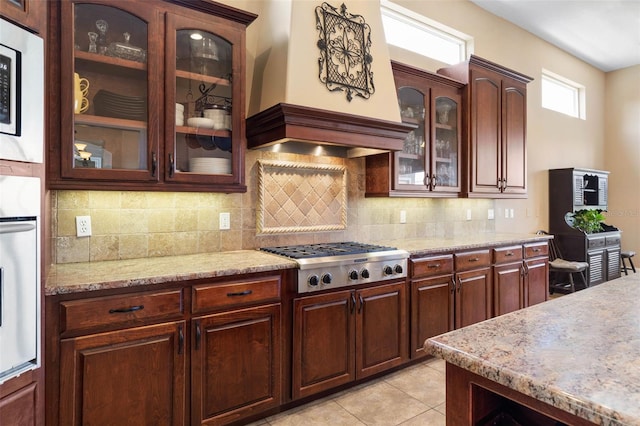 kitchen featuring light stone counters, tasteful backsplash, glass insert cabinets, custom exhaust hood, and stainless steel appliances