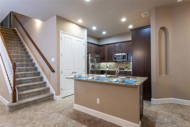 kitchen featuring light stone counters, stainless steel appliances, tasteful backsplash, an island with sink, and dark brown cabinets