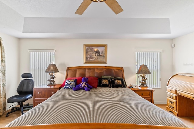 bedroom with light wood-type flooring, a tray ceiling, baseboards, and a ceiling fan