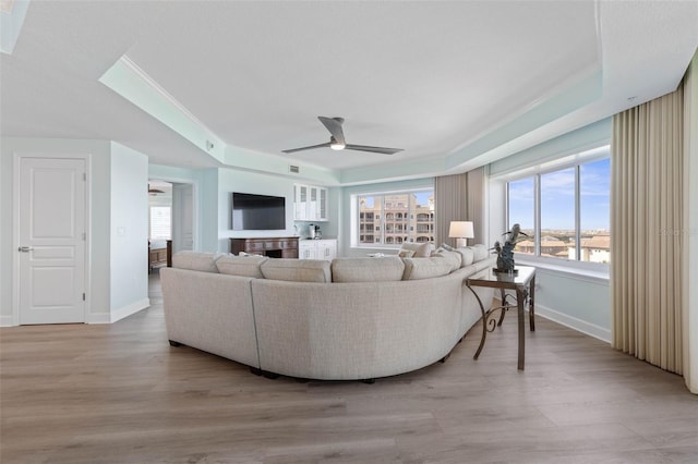 living room featuring a ceiling fan, baseboards, light wood finished floors, ornamental molding, and a raised ceiling