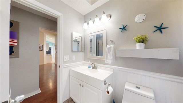 half bathroom with vanity, wood finished floors, a wainscoted wall, a textured ceiling, and toilet