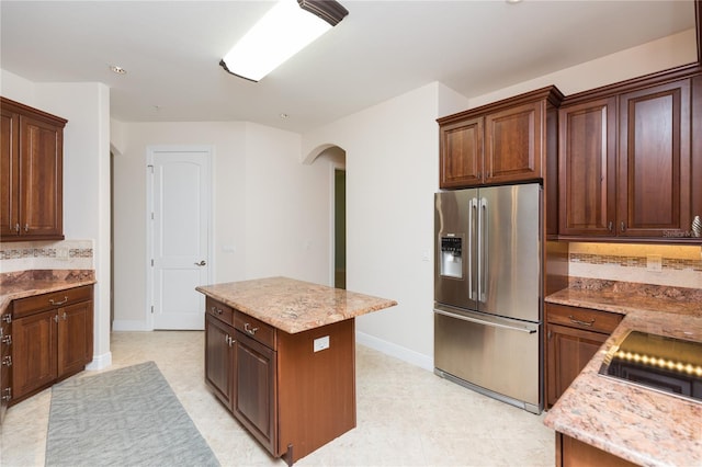 kitchen featuring stainless steel refrigerator with ice dispenser, a kitchen island, arched walkways, decorative backsplash, and light stone countertops