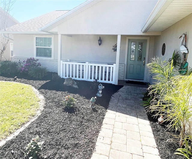 doorway to property featuring a shingled roof, a porch, brick siding, and stucco siding