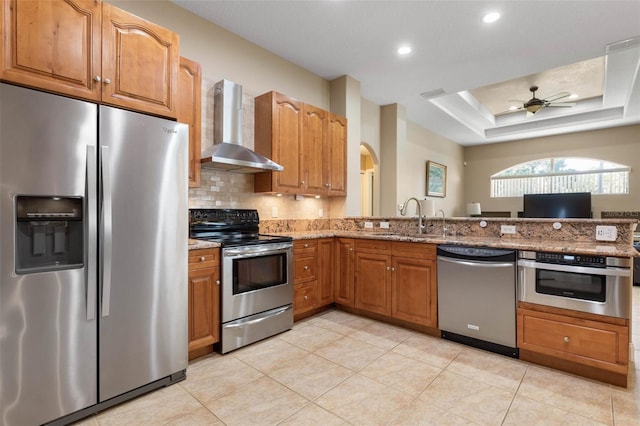 kitchen featuring a sink, stainless steel appliances, wall chimney range hood, a raised ceiling, and light stone countertops