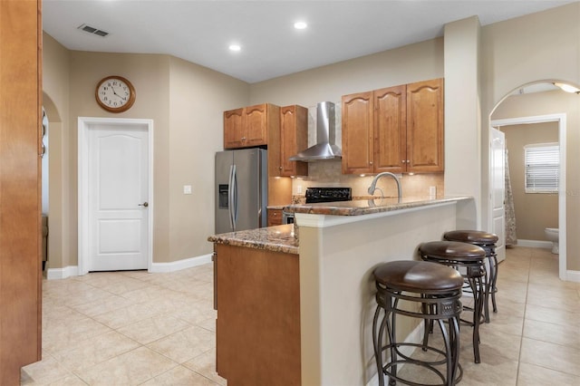 kitchen featuring a breakfast bar, range with electric stovetop, stainless steel refrigerator with ice dispenser, arched walkways, and wall chimney exhaust hood