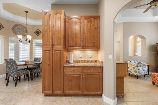 kitchen with light tile patterned floors, light stone countertops, a raised ceiling, ceiling fan with notable chandelier, and tasteful backsplash