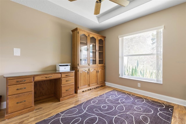 office featuring ceiling fan, a tray ceiling, baseboards, and light wood-type flooring