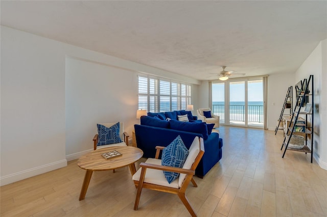 living room featuring light wood-type flooring, a water view, a wall of windows, baseboards, and ceiling fan