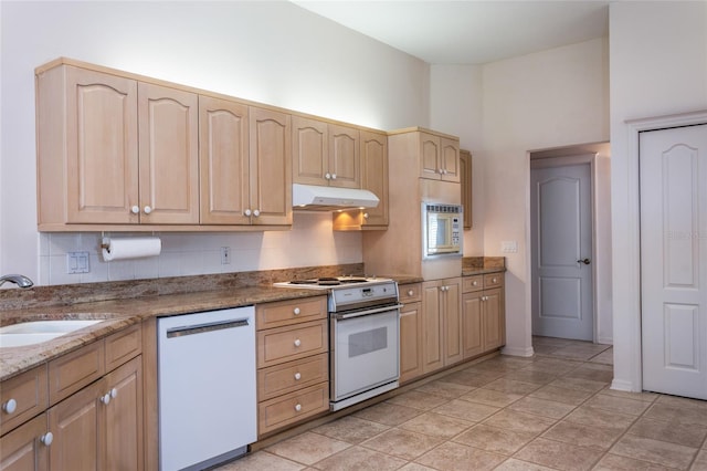 kitchen featuring white appliances, light stone counters, light brown cabinets, a sink, and under cabinet range hood