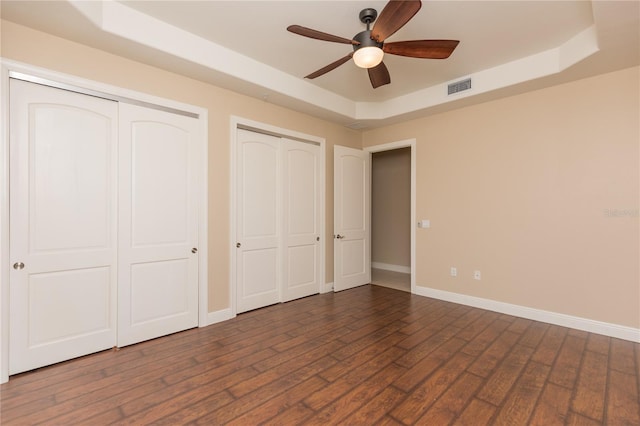 unfurnished bedroom featuring visible vents, two closets, a tray ceiling, baseboards, and dark wood-style flooring